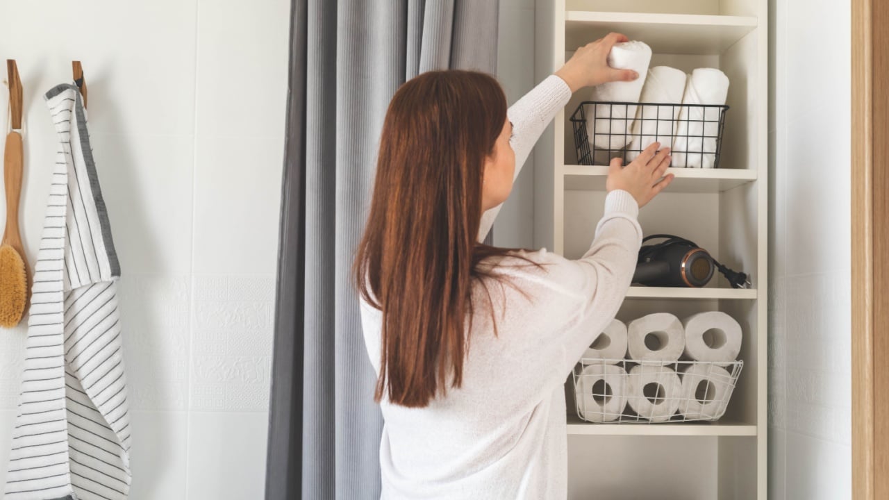 Happy young woman is neatly placing rolled up white towels into a metal mesh basket near bath sink in bathroom. Bath towels storage solution and space saving concept. Personal hygiene and care.