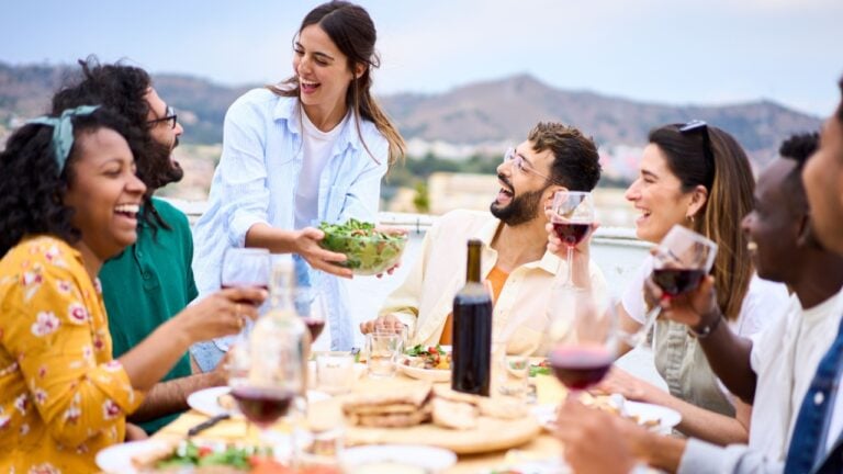 Young smiling Caucasian woman putting plate with food on table at celebration with happy friends on rooftop. Group of cheerful multiracial friends gathered for barbecue party on outdoor terrace.