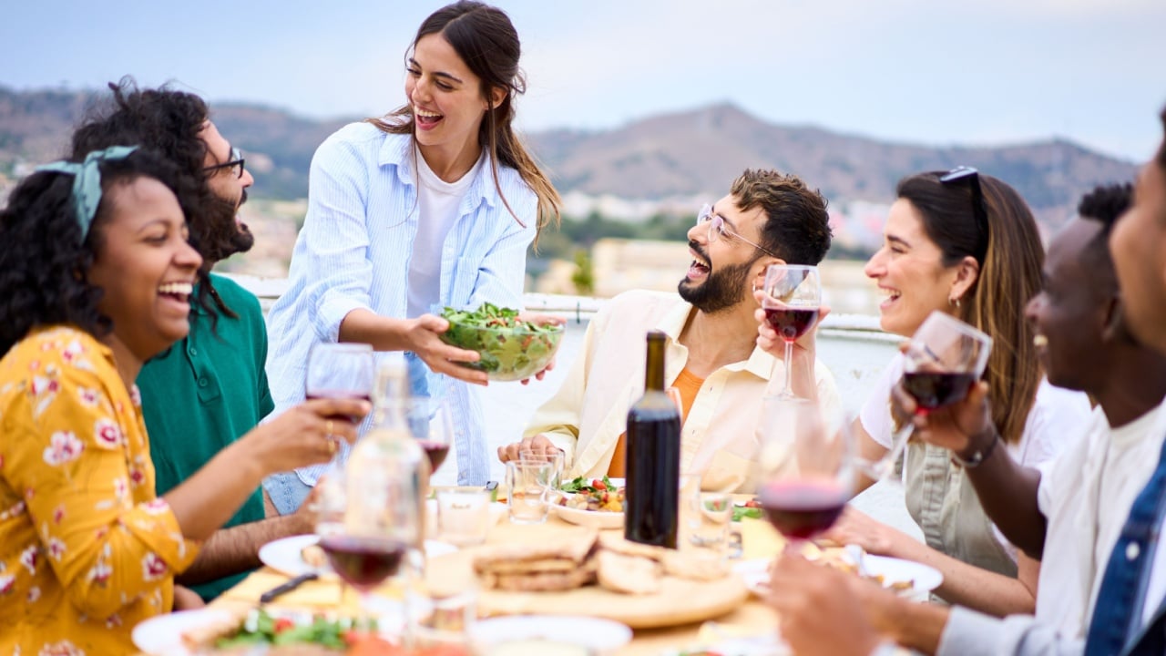 Young smiling Caucasian woman putting plate with food on table at celebration with happy friends on rooftop. Group of cheerful multiracial friends gathered for barbecue party on outdoor terrace.