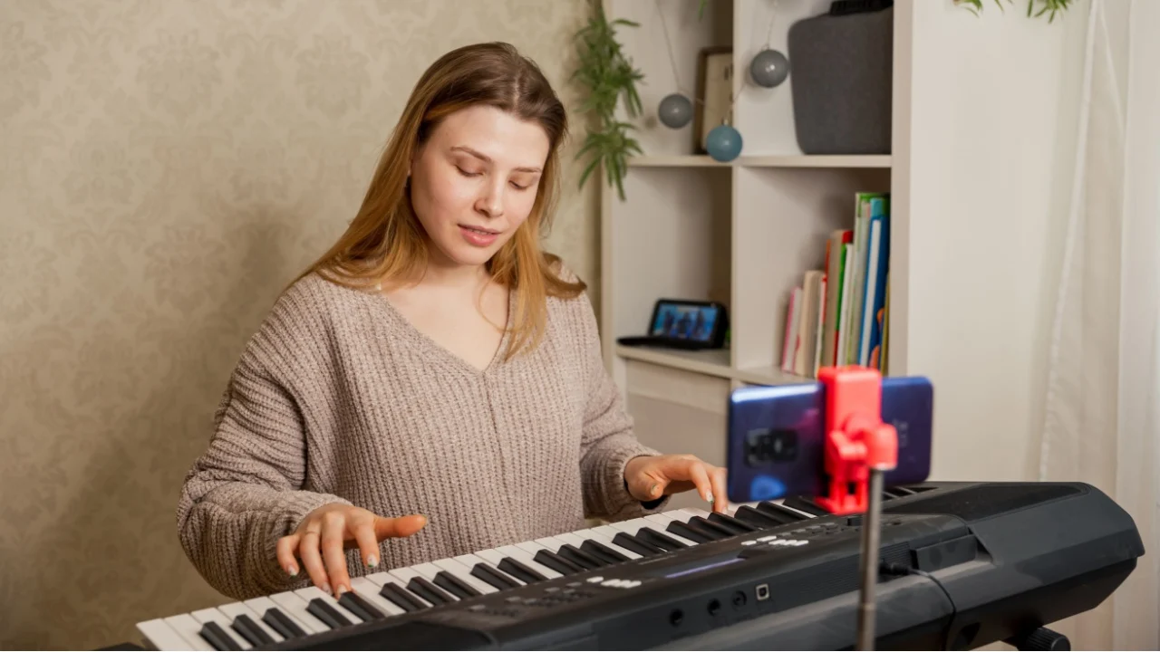 Music teacher turning on smartphone for online lesson, while sitting at table with synthesizer at home. Woman learns music, playing piano online at home interior. Female play piano synthesizer