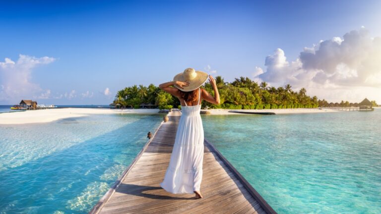 Back view of a elegant woman in white dress and hat walks down a pier towards a tropical island in the Maldives