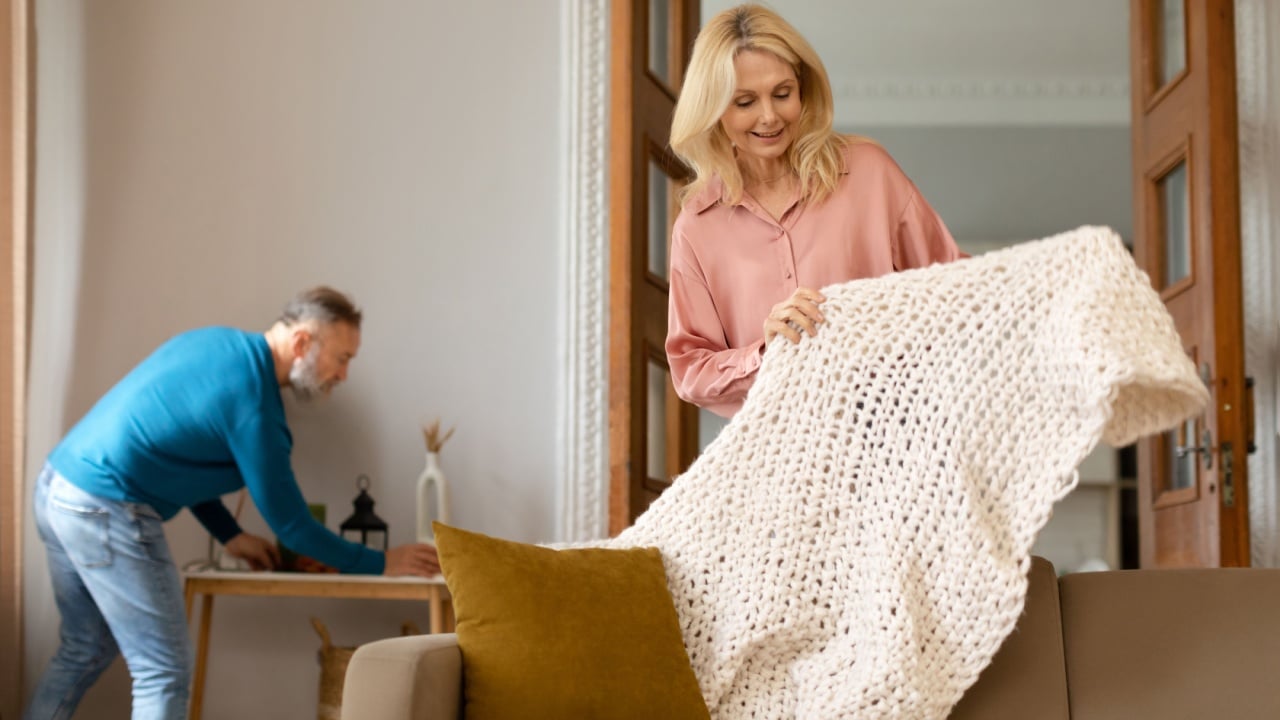 Domestic Comfort. Happy Senior Couple Tidying And Cleaning Cozy Living Room Indoor, Woman Covering Sofa With New Knitted Blanket While Her Husband Dusting Furniture. Housework Chores