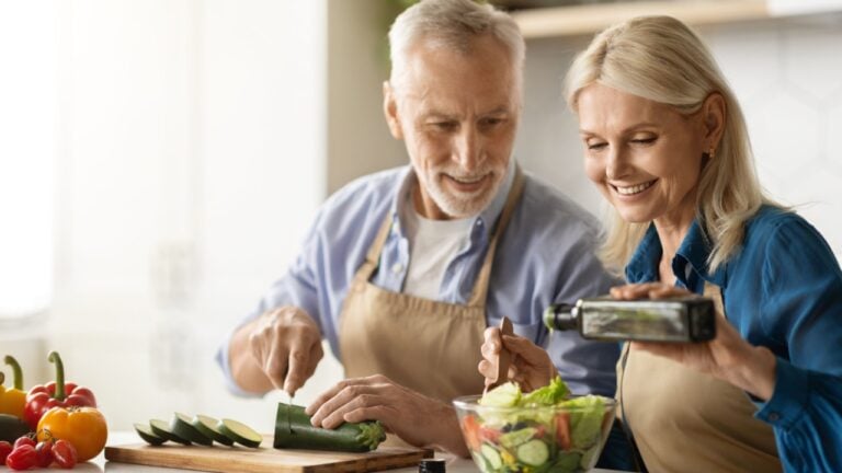Happy Senior Couple Preparing Tasty Lunch Together In Kitchen, Smiling Mature Man And Woman Cooking Healthy Vegetarian Meal At Home, Woman Adding Oil To Fresh Vegetable Salad.