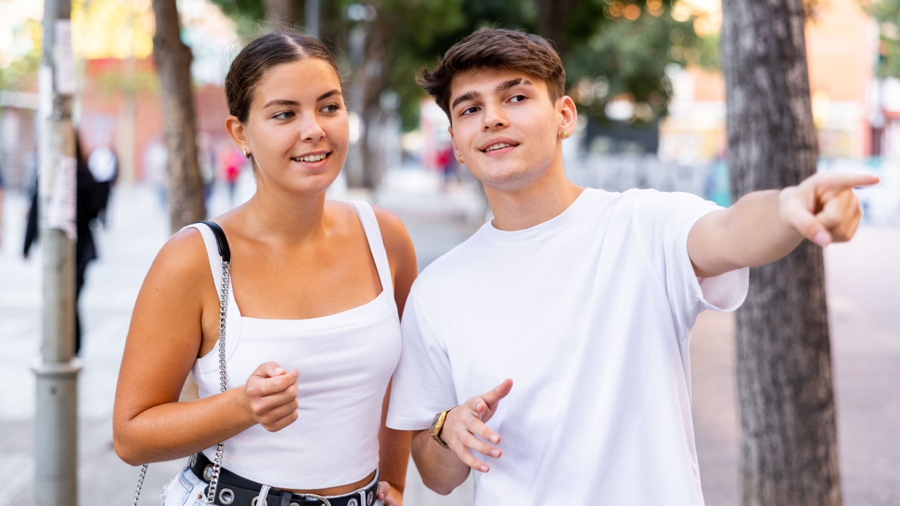 Young man and woman walking through city streets together, pointing finger to show direction.