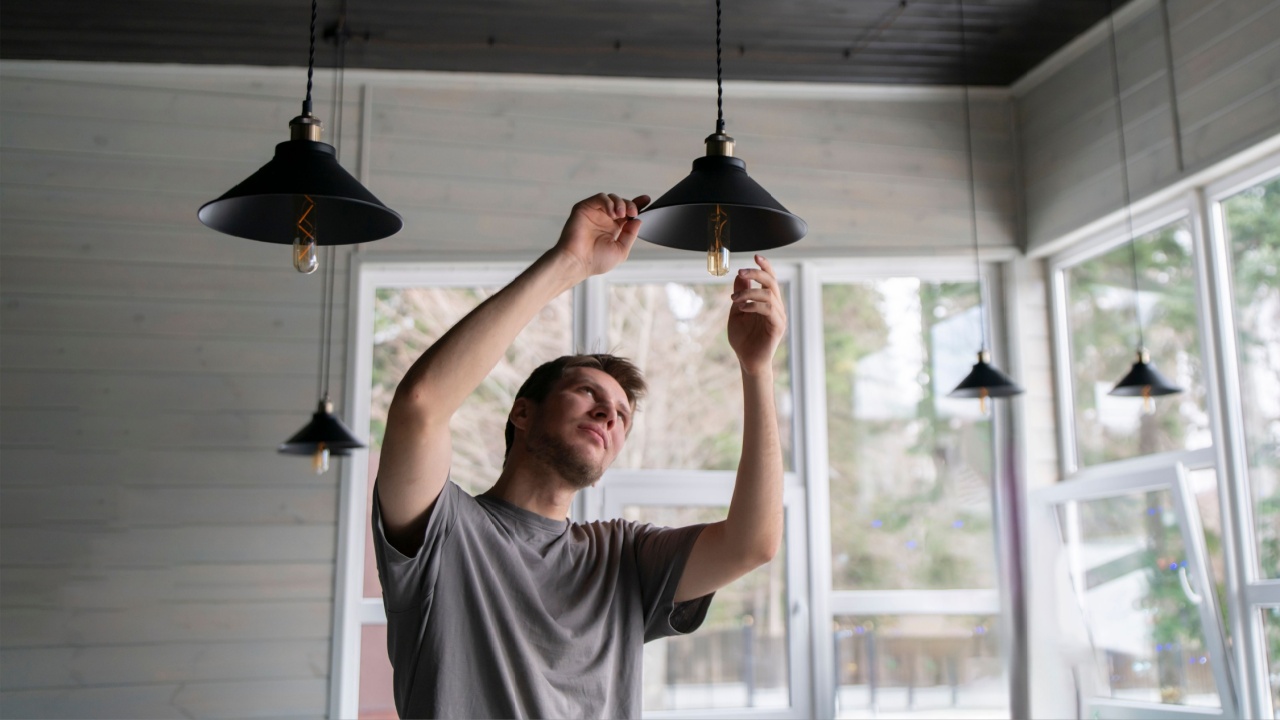 A man is captured in the act of changing a light bulb in a hanging ceiling light fixture, with natural daylight illuminating the scene.