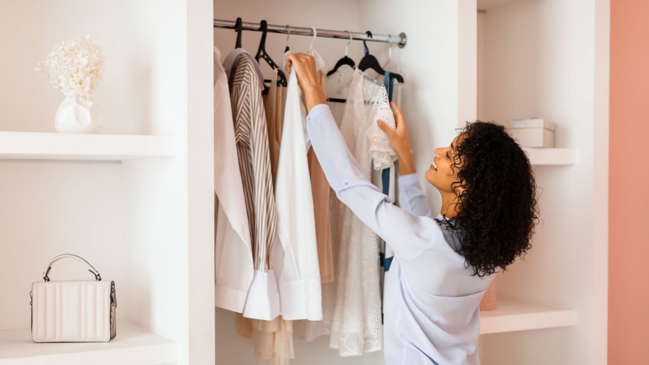 Woman with curly hair is selecting white blouse from her fashionable wardrobe in a room with chic, minimalist decor and soft lighting