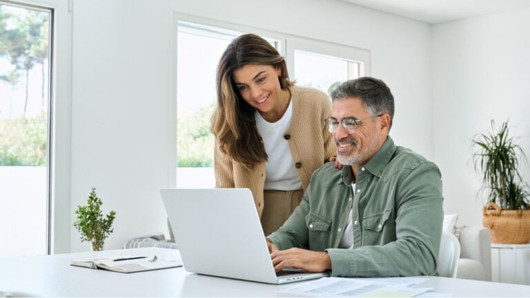 Smiling middle aged senior man working on computer sitting at table with wife standing nearby in living room. Happy mature older couple using laptop technology at home. Authentic candid shot.