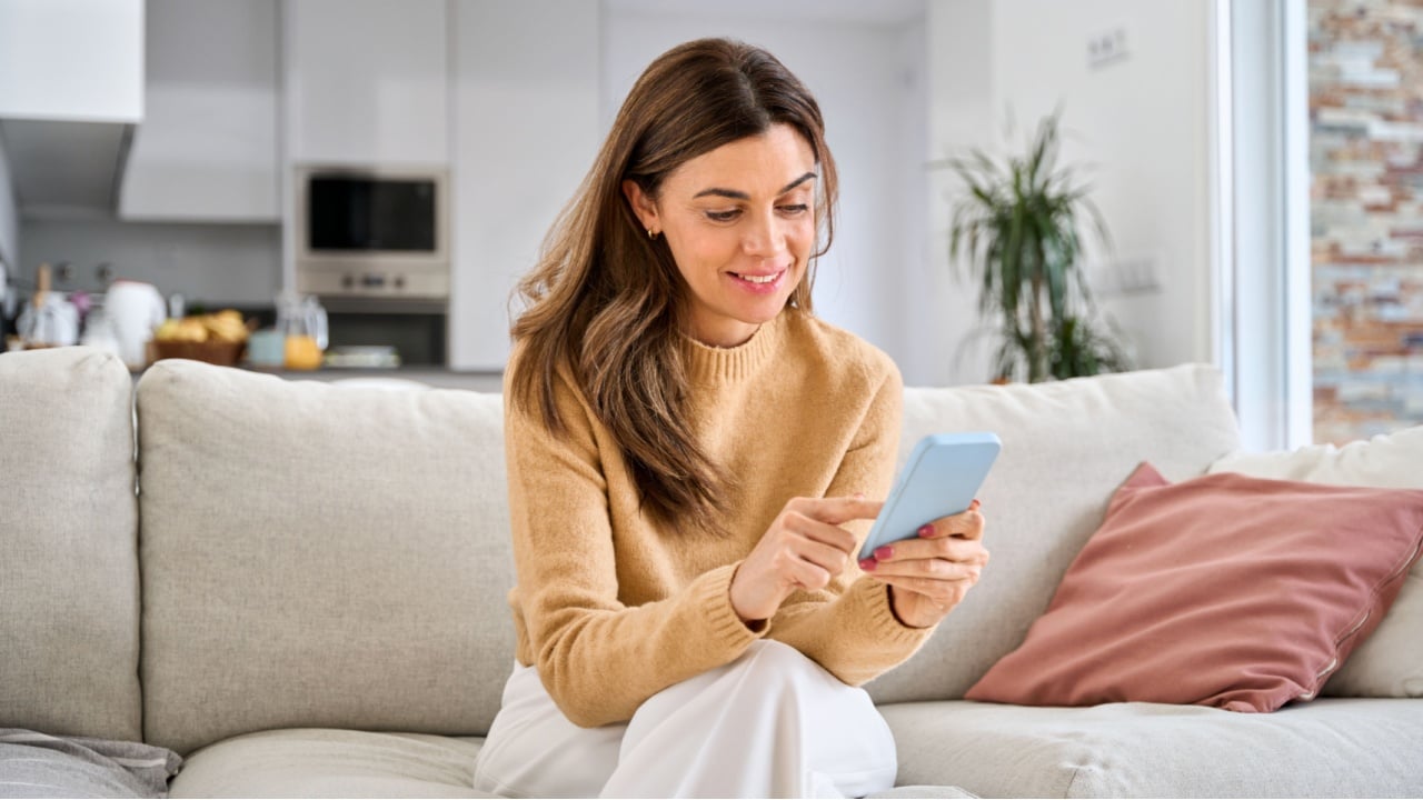 Happy mature older woman using smartphone sitting on couch at home. Smiling middle aged woman looking at cell phone messaging or doing ecommerce mobile shopping relaxing on sofa in living room.