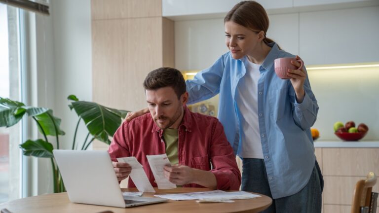 Thoughtful husband sitting at table with laptop, holding bills, counting payments, savings, analysing income, expenses. Wife interested in family budget planning, looking at display while drinking tea