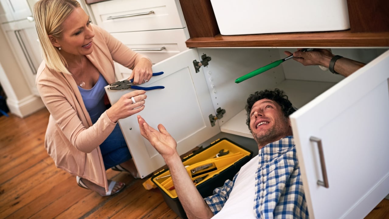 Lady, help and handyman with tools for fixing home basin, drain or broken interior sewer. Plumber, working and woman watching male person repair for burst pipe, sink leaks and kitchen maintenance