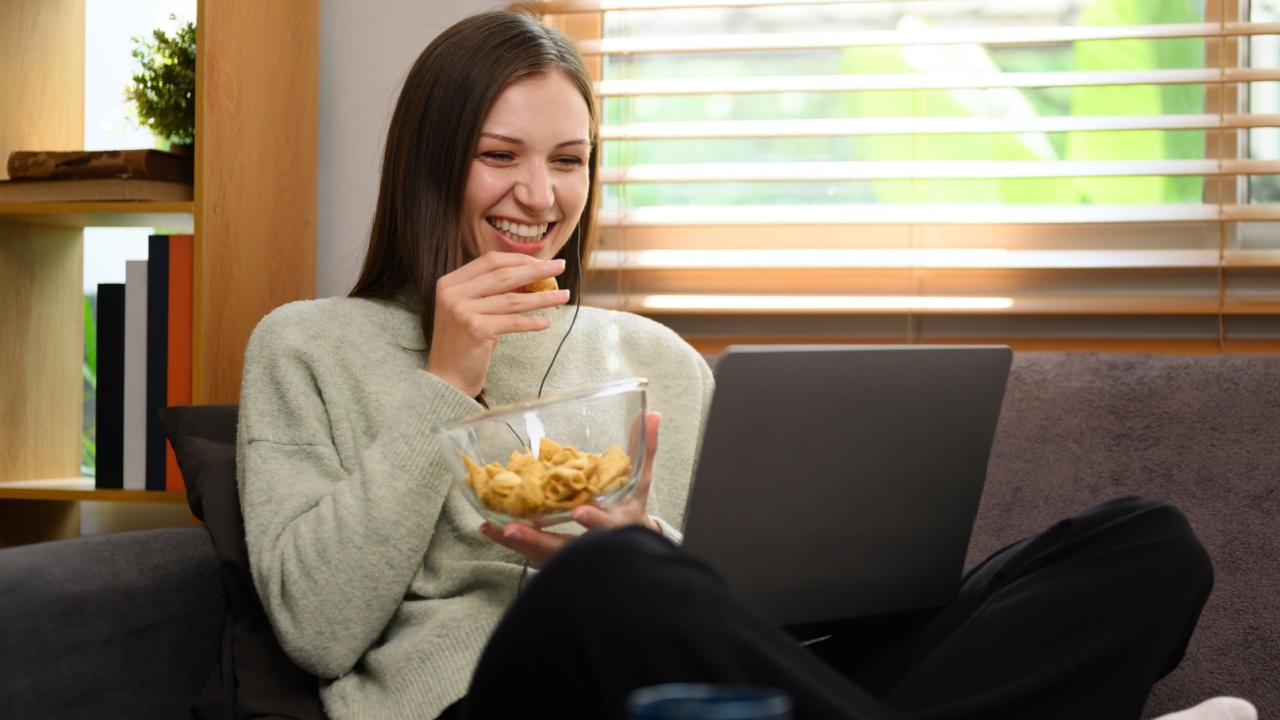 Happy young woman eating cookies and watching a movie on a laptop at home
