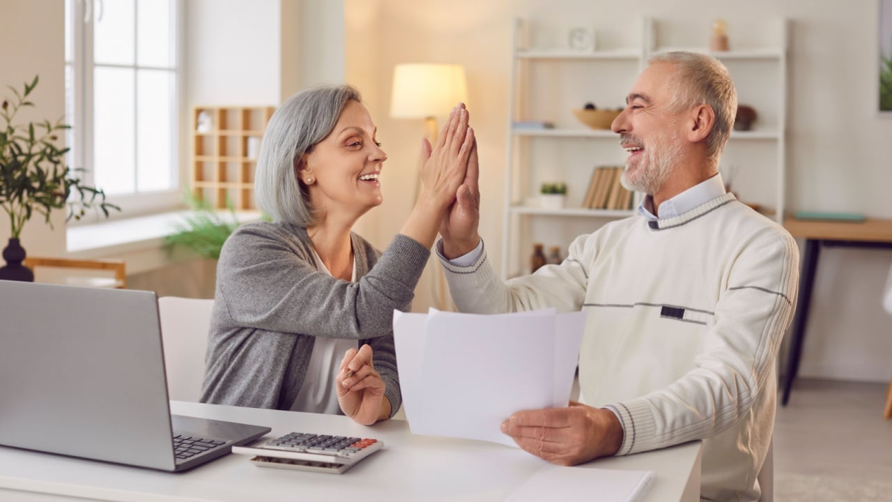 Senior couple sitting at the table with laptop and bills giving high five each other calculating finances or taxes at home. Elderly retired man and woman rejoicing income and profit on pension.