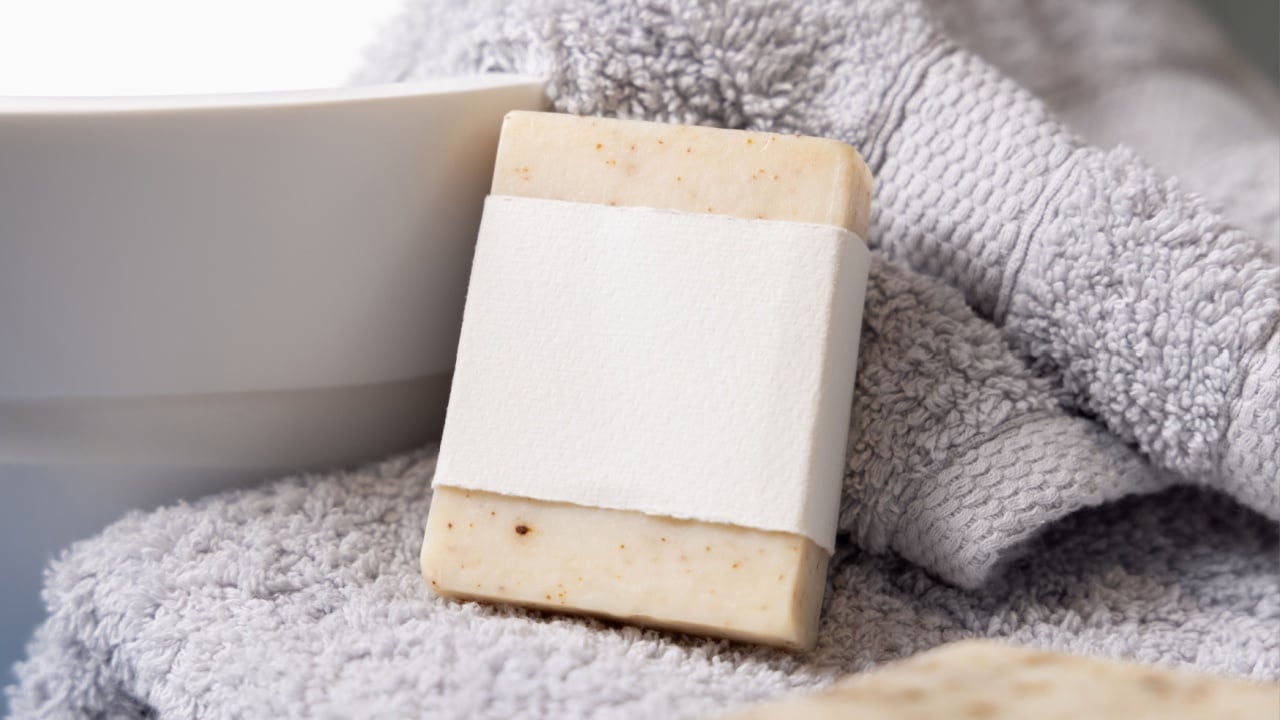 Beige soap bar with horizontal blank label on light grey towels near vessel sink in bathroom.