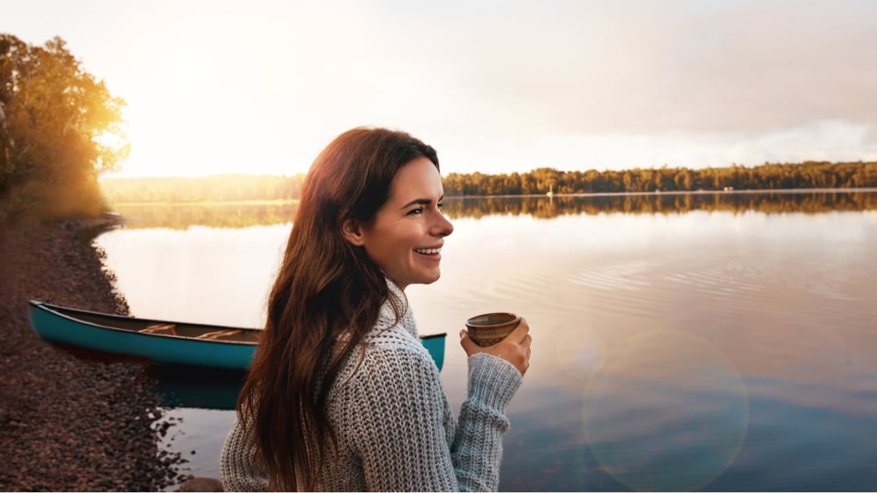 Nature, river and woman with coffee to enjoy, sunrise and outdoor with smile in vacation and morning. Break, relax and girl with joy for inspiration in countryside of Finland with peace by lake