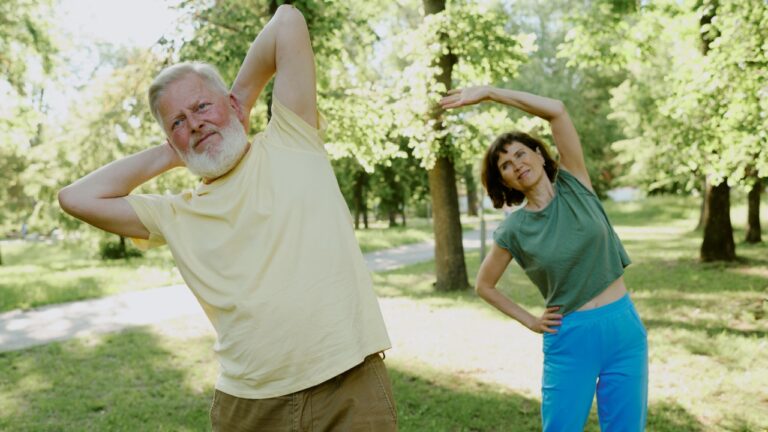 Senior silver-haired man and brunette woman dressed in cotton sportswear doing back exercise in park