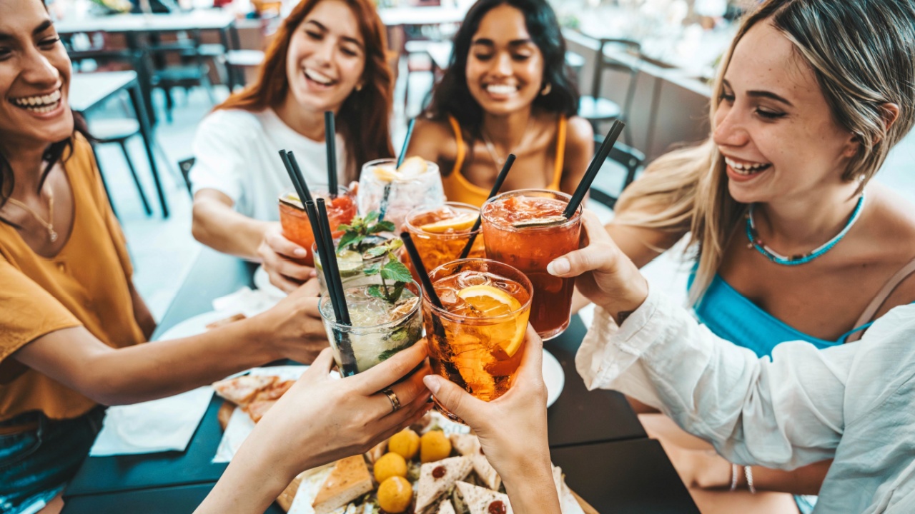 Happy female people toasting cocktail glasses sitting at bar table - Cheerful friends enjoying happy hour at pub restaurant balcony - Food and beverage life style concept