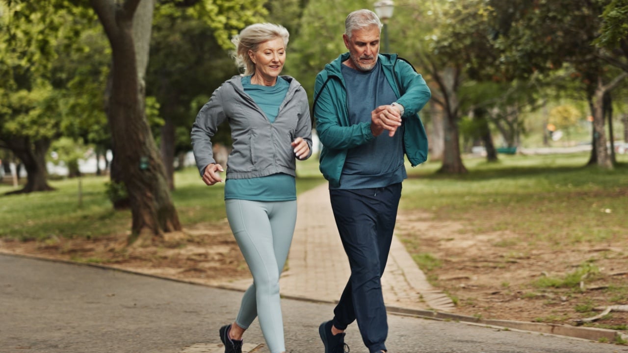 Senior couple, running and smart watch in park with check for speed, steps or hear rate for fitness. People, man and woman with timer, stopwatch and exercise in nature, path and training in Germany