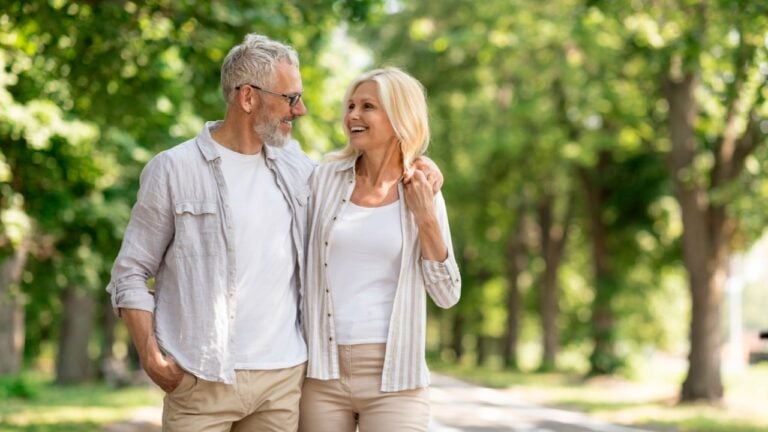 Romantic Senior Couple Walking Outdoors In Summer Park And Embracing, Loving Happy Mature Spouses Hugging And Smiling To Each Other, Cheerful Husband And Wife Enjoying Outside Walk.