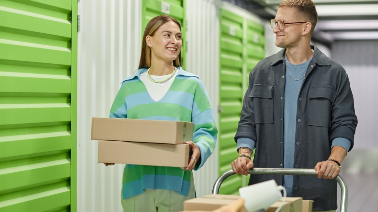 Medium shot of cheerful woman carrying cardboard boxes walking along self storage warehouse hallway with helping boyfriend pushing dolly cart during apartment decluttering