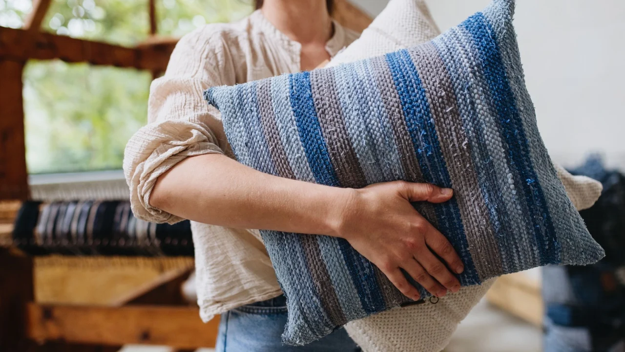 Young businesswoman next to wooden loom, holding pillows with woven covers made from recycled fabrics. Upcycling in business and circular economy.