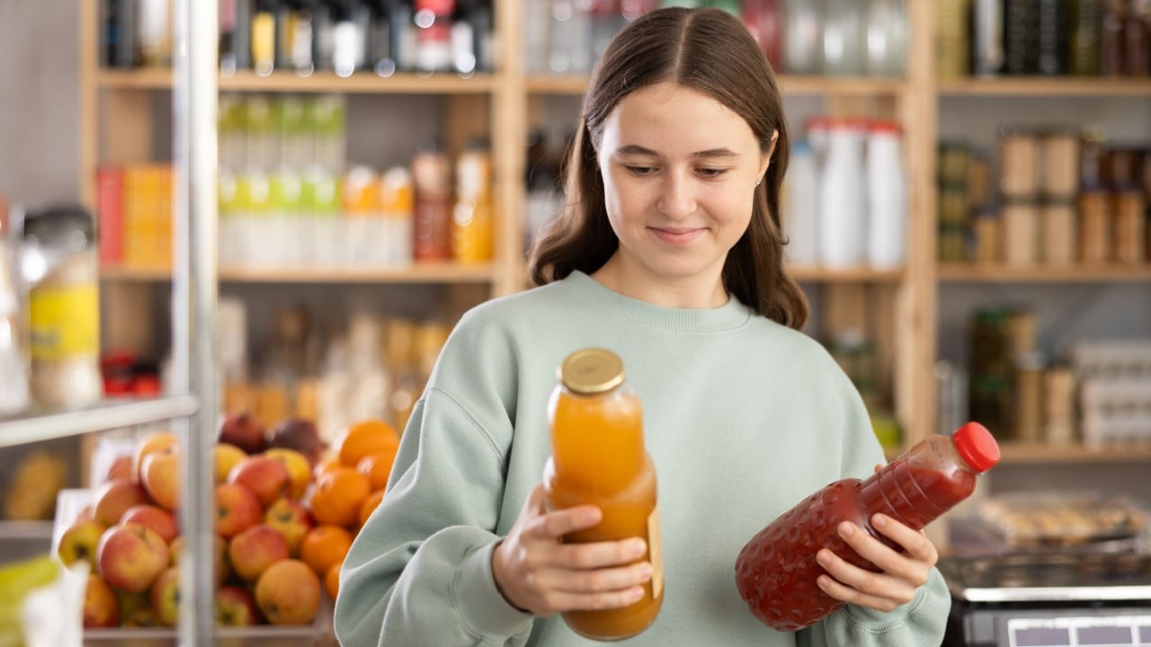 Teen girl hold packages bottles and choose consider juice in store. Market visitor choose consider make choice bottle fruit drink. Buyer pick up set of groceries for week.