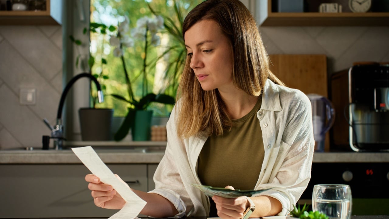 Stressed woman reviewing bills at home. Female organizing receipts, sitting at kitchen table with laptop and paper documents. Concept of budgeting and managing personal finances