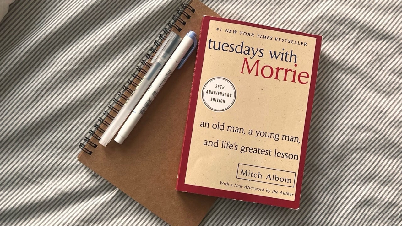 A copy of Tuesdays with Morrie rests on a beige notebook with a pen beside it on a soft, striped bedspread. This setup invites contemplation and deep thought.
