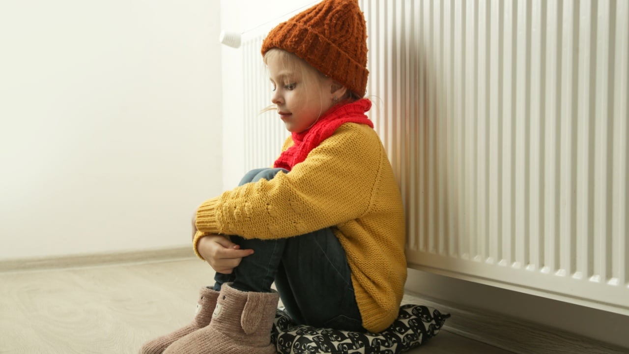 Little girl in woolen winter clothes, near the radiator of the home heater trying to keep warm. Restrictions and savings of gas and electricity during energy or economic crisis, rising prices.