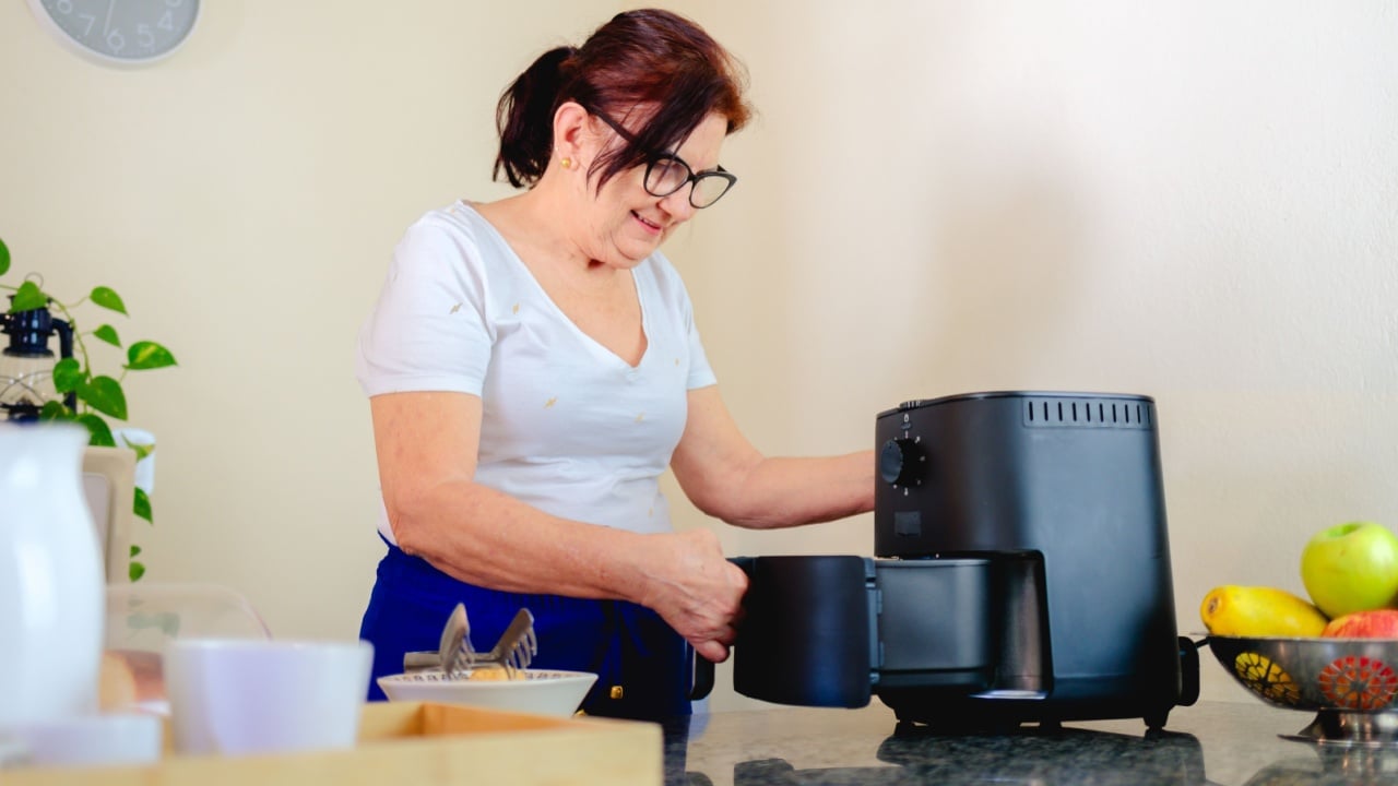 Smiling mature woman using an air fryer on the countertop of her kitchen. Healthy cooking concept with modern appliances. Cozy home scene in a typical Brazilian house