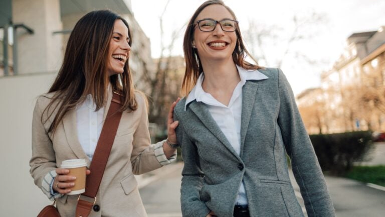 Two cheerful businesswomen are walking down the street, enjoying a coffee break and engaging in a friendly conversation, showcasing camaraderie and work life balance