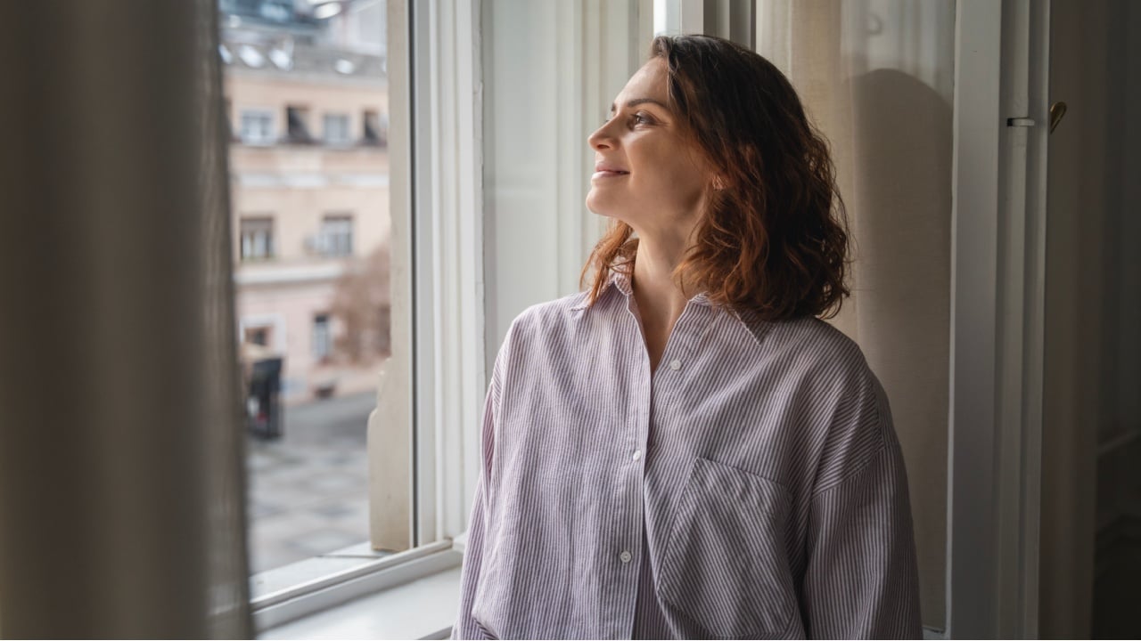 Young relaxed woman standing at window in apartment overlooking european city