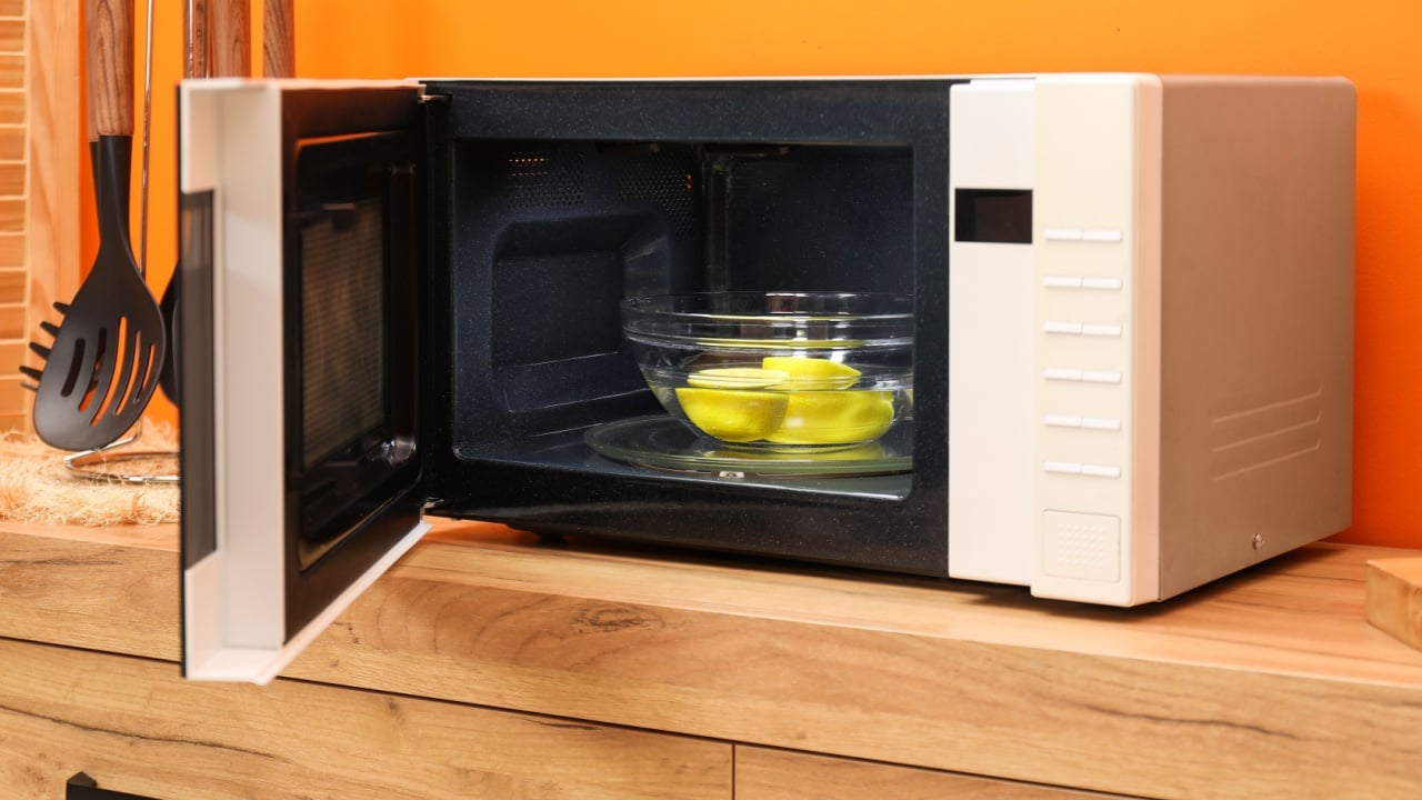 Open microwave oven with bowl of water and lemons on wooden cabinet near orange wall indoors