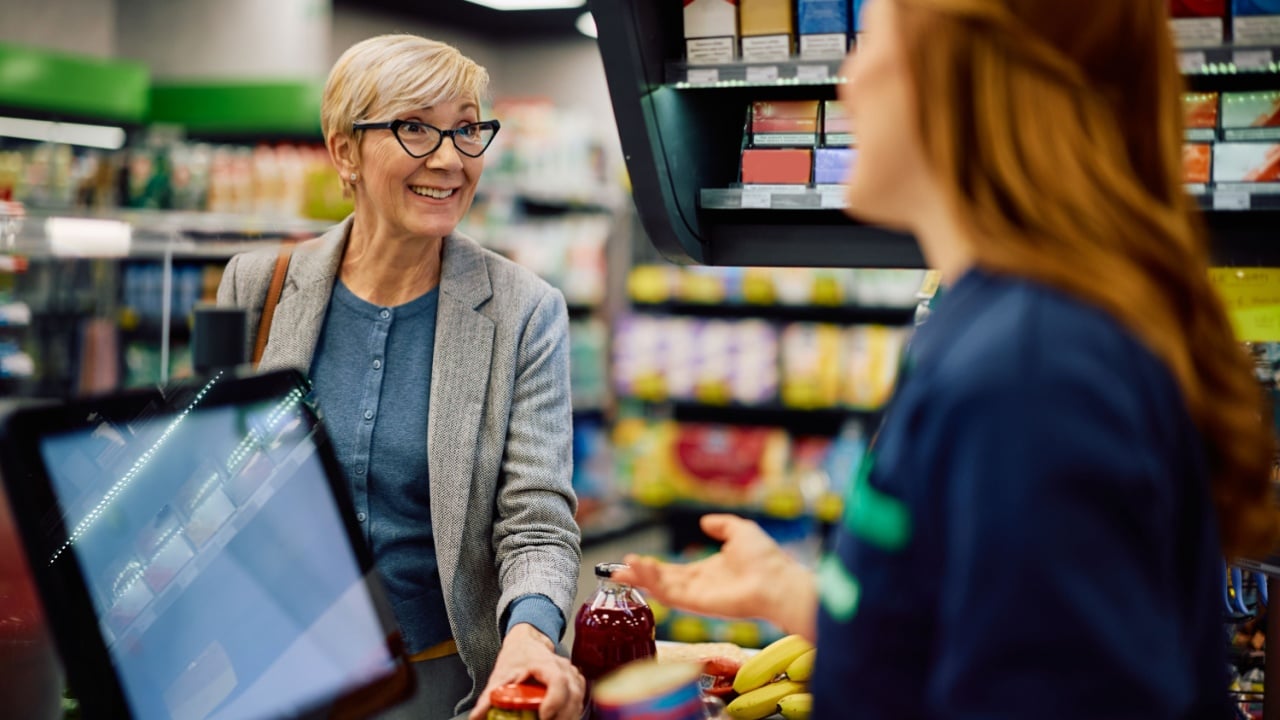 Happy senior woman putting groceries on checkout counter while talking to cashier at supermarket.
