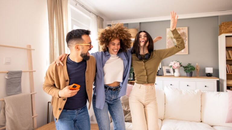 Wide-angle shot of three energetic young adults, including two women and one man, dancing joyfully on a white couch in a naturally lit, contemporary living room setting.