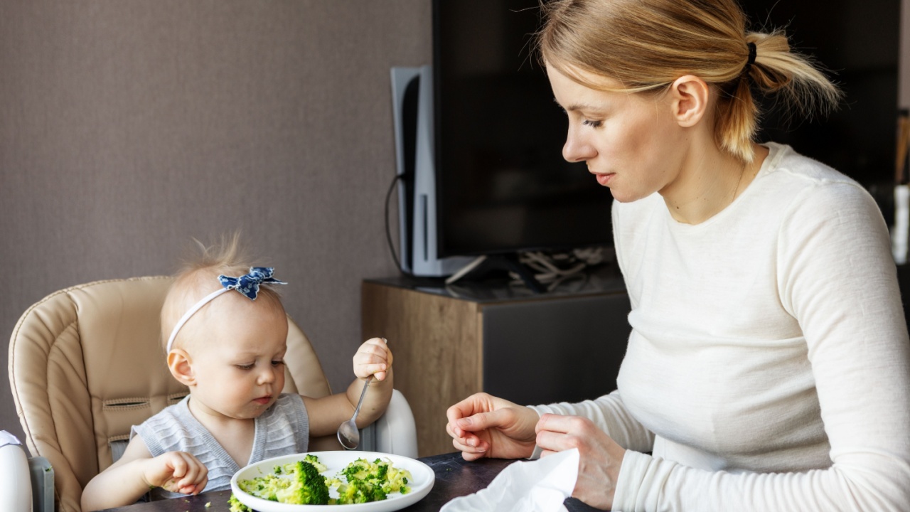 Little baby eating healthy broccoli with spoon while sitting in high chair, mother gently helping and supporting child during mealtime. Real life parenting, nutrition, bonding and family care indoors