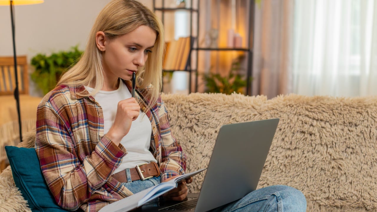 Young adult woman writes in notebook while focusing on laptop screen on home sofa, calculating expenses. Blonde girl manages financial records with care, staying organized and concentrated on task.