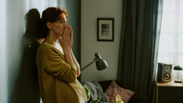 Woman standing by wall in living room, crying. Room decorated with lamp, framed photo, and few plants, creating a serene and thoughtful atmosphere