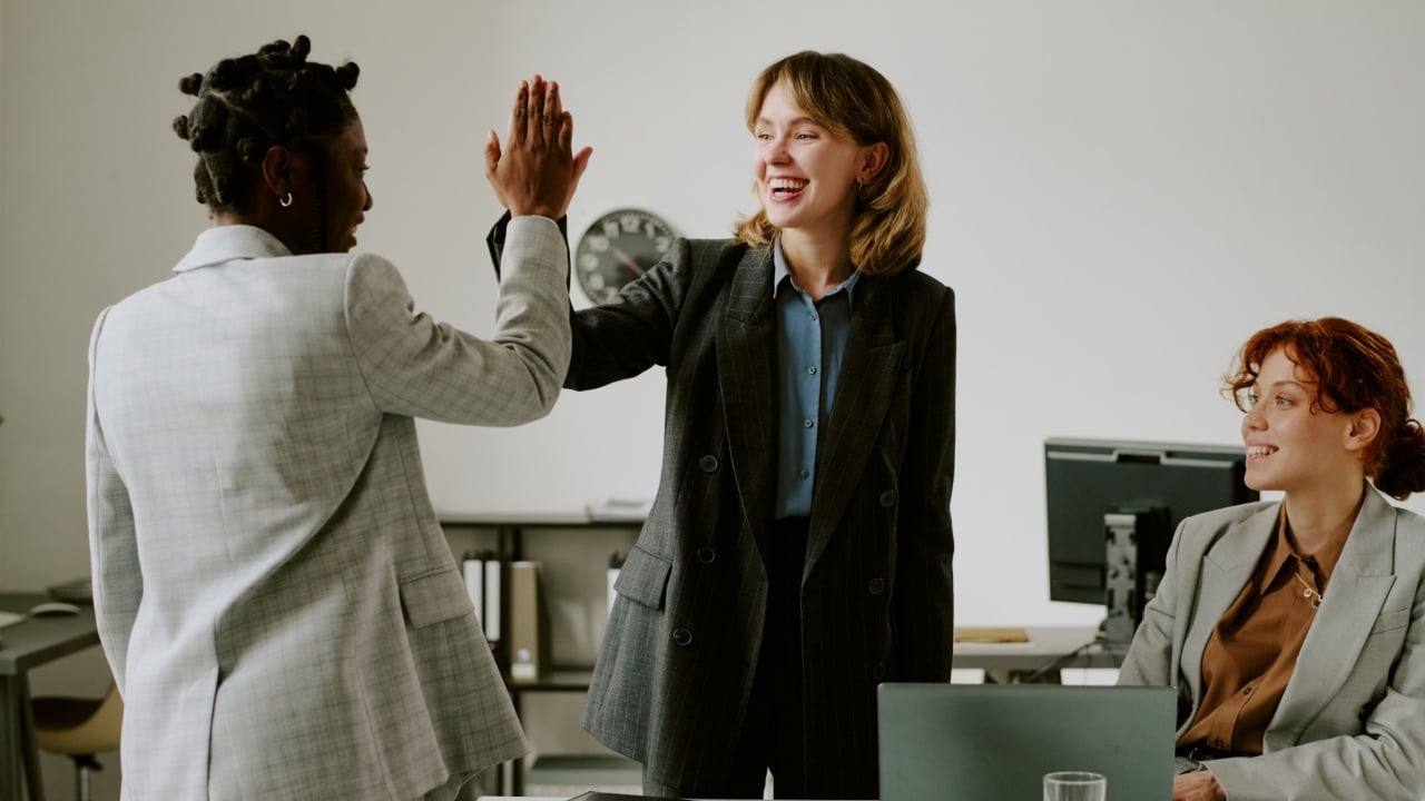 Group of multicultural businesswomen celebrating successful achievement in modern office setting, showing excitement and camaraderie during work