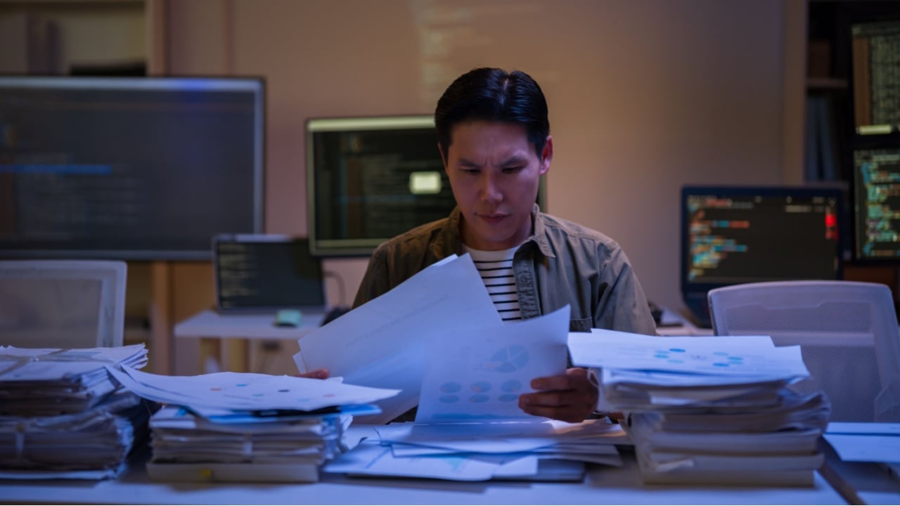 Asian man working late at night with busy paperwork piled up on her desk, stressed out from not being able to finish her work in the office