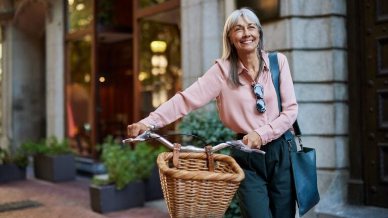 Happy senior woman in casual smiling while holding her bicycle. Older lady walking outside with her bike on an urban scene, copy space. Mature woman enjoying a peaceful walk with cycle in town.