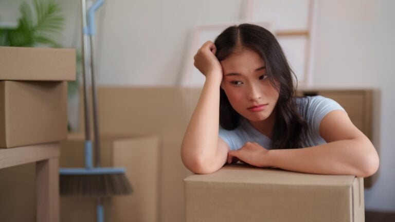 Young adult Asian female experiences moving day fatigue surrounded by relocation packing boxes ready for transport or unpacking in new apartment residence
