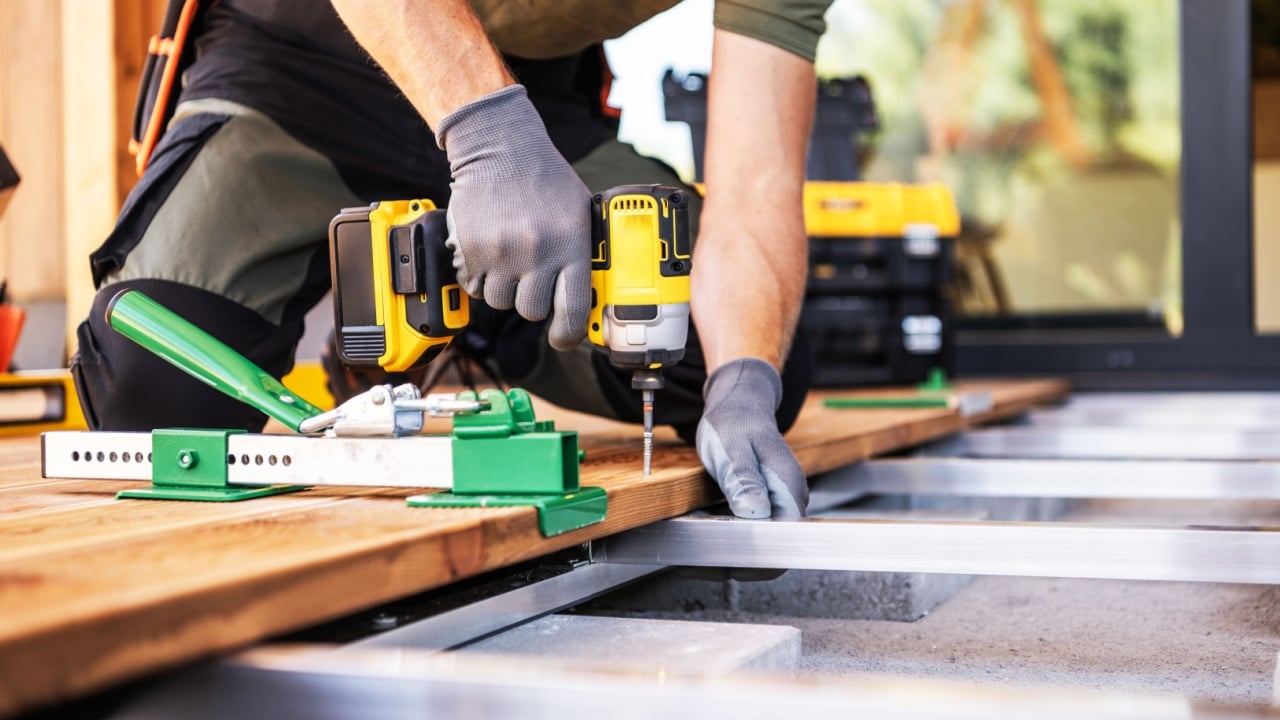 A skilled craftsman uses a drill to secure wooden planks on a deck frame while wearing gloves. Tools and building materials are organized nearby in a home setting.