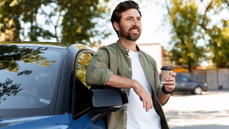 Smiling businessman drinking takeaway coffee while leaning on his electric car in a parking lot
