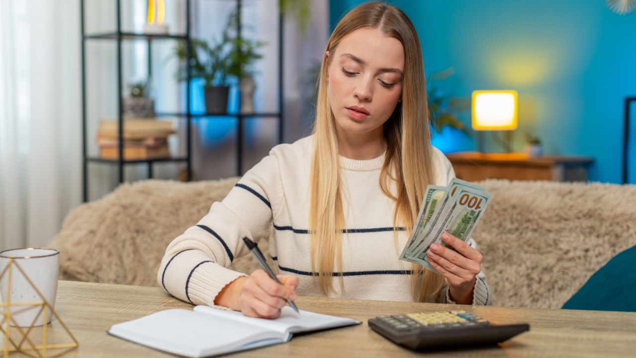 Caucasian woman at home on table counting cash, using calculator, writing in notebook, feeling very confident about salary. Girl appears happy, satisfied, enjoying income, money management, plans