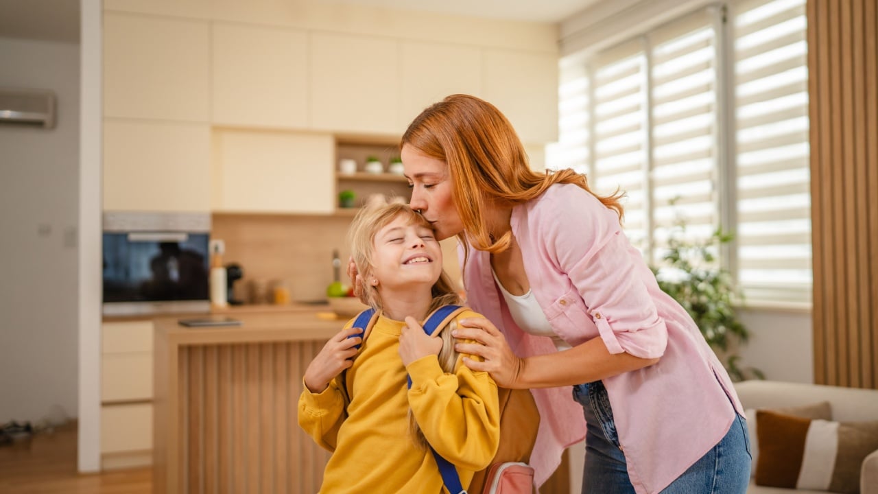 Loving mother is kissing her happy daughter on the forehead before school, showing affection and support in their modern and bright kitchen