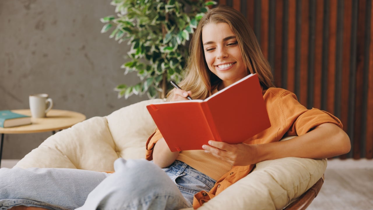 Young happy woman wear orange casual clothes write down memories in notebook diary sits in chair stay at home hotel flat rest relax spend free spare time in living room indoor.