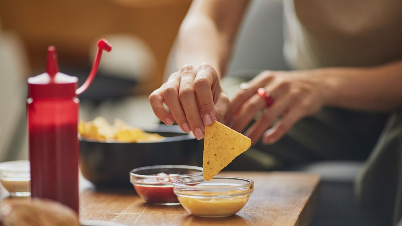 Caucasian young adult woman dipping tortilla chip into cheese sauce while sitting at table with snacks, hand visible reaching toward bowl, ketchup bottle and food in foreground