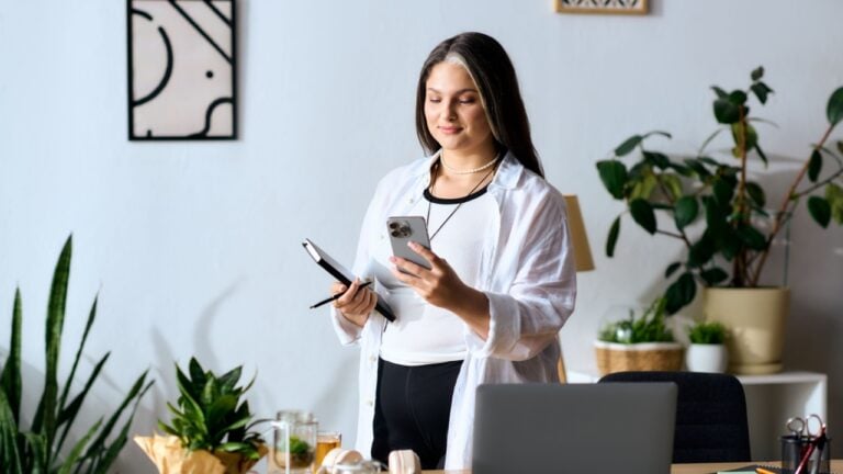 A woman with Waardenburg syndrome stands in her home office, reviewing notes and using her phone.
