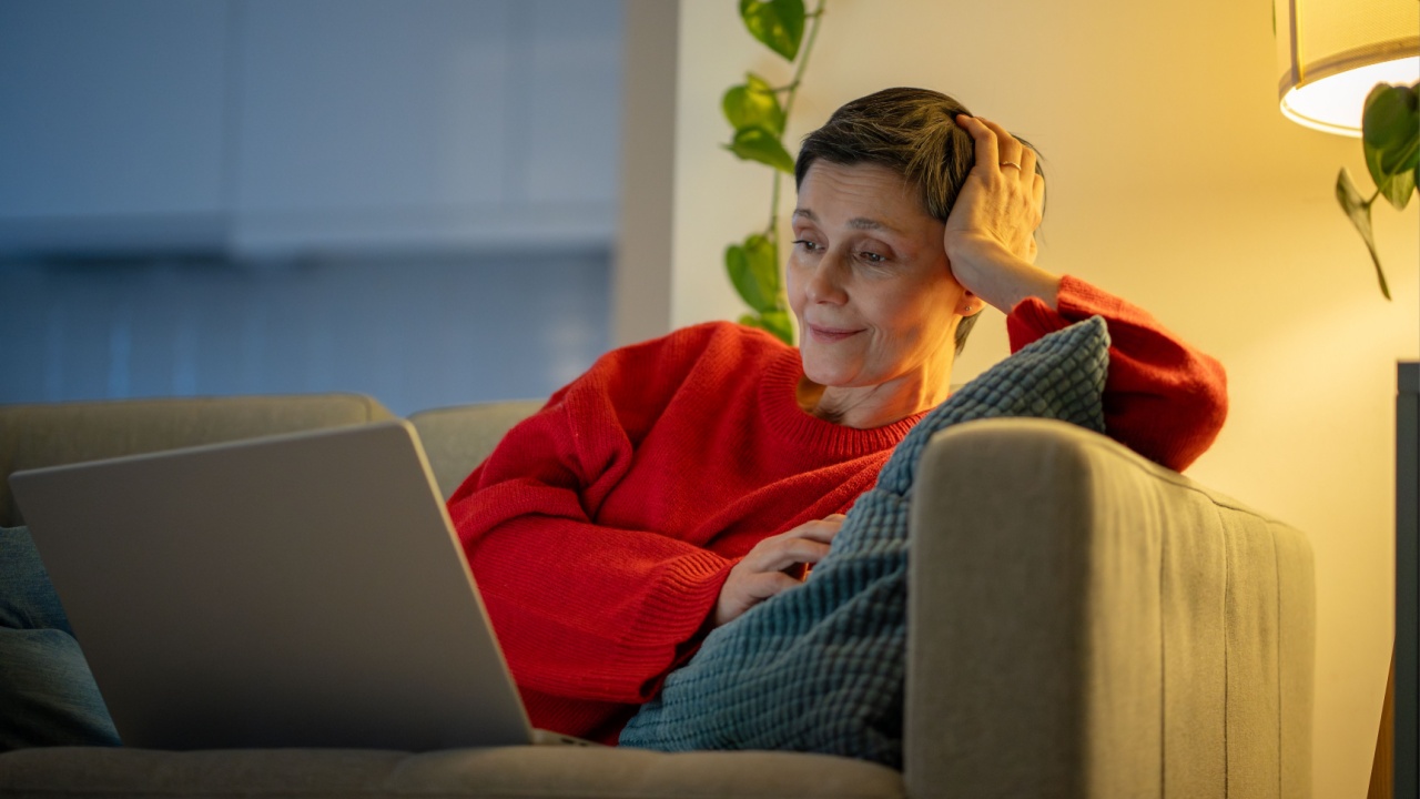 Middle aged smiling woman laying on sofa in living room at evening and watching movie on laptop. Elderly female relaxing on couch and watching video on computer, relaxing after work. Cozy leisure time