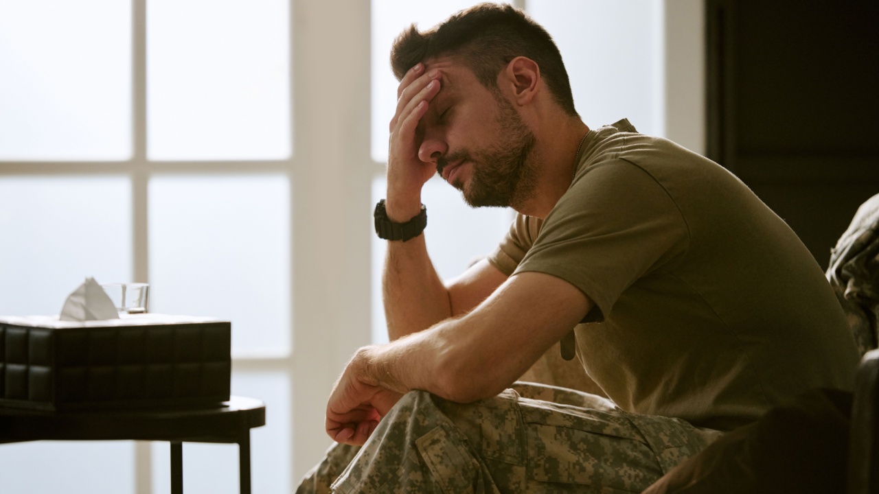 Middle aged Caucasian man sitting with hand on forehead, showing signs of emotional distress wearing military uniform in bright room suggesting psychological struggle or mental health issue